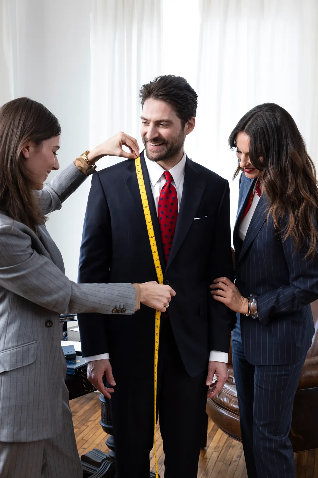 A tailor measuring a client in a navy suit with a yellow tape measure