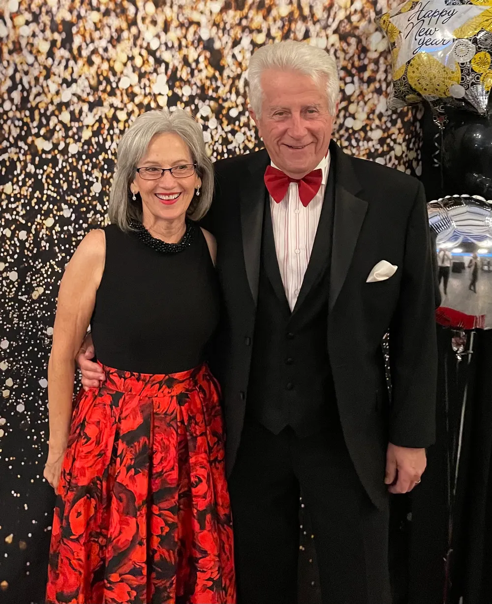 A man in a custom black tuxedo and three-button vest with a red bow tie, standing with his wife at a black-tie New Year party