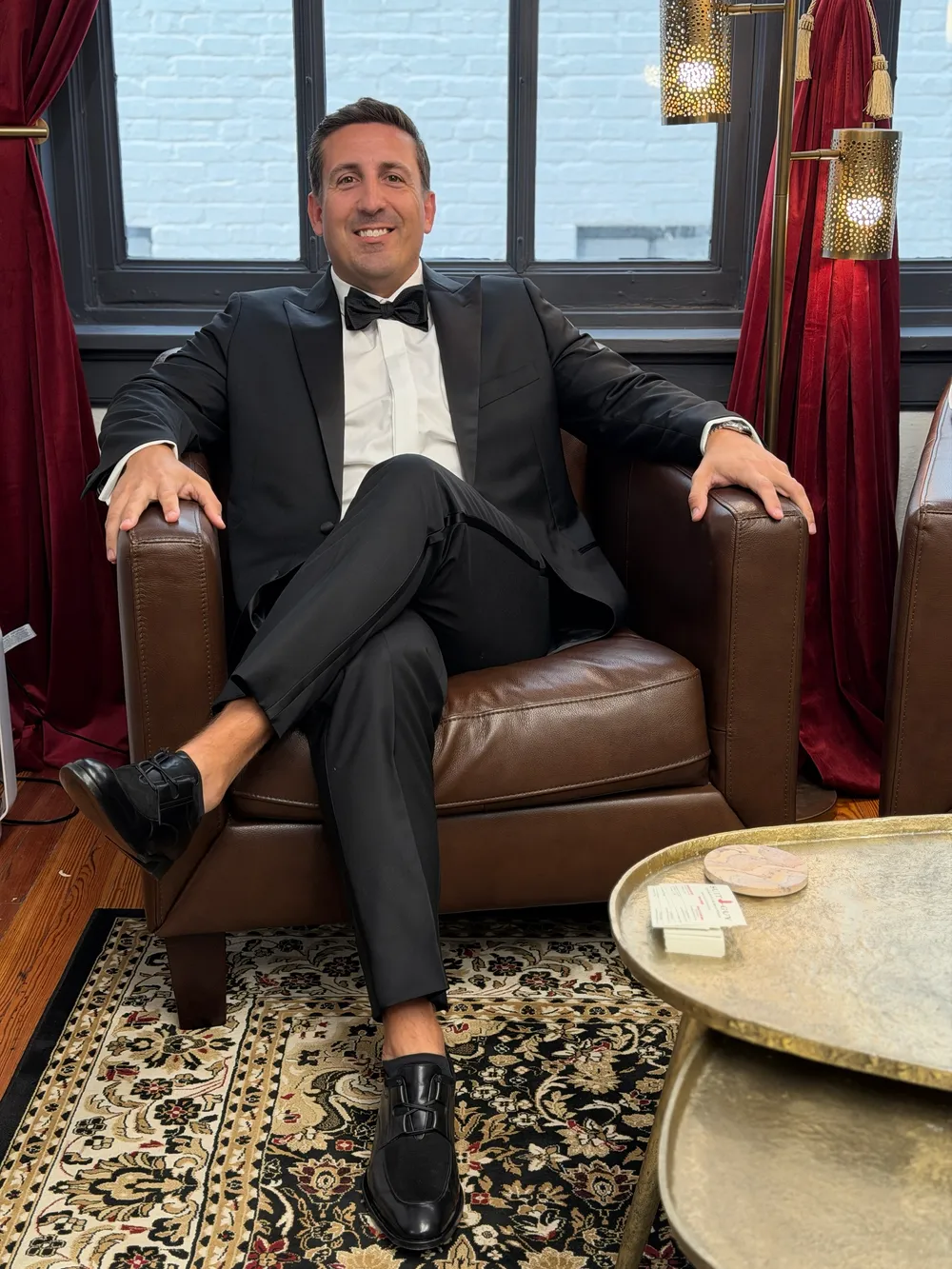A groom in a custom black peak-lapel tuxedo and bow tie, relaxed in a leather armchair