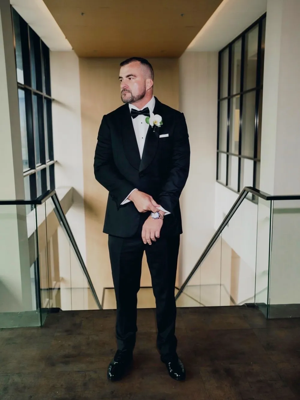 A groom in a custom shawl-collar tuxedo adjusting his cuff on a modern staircase